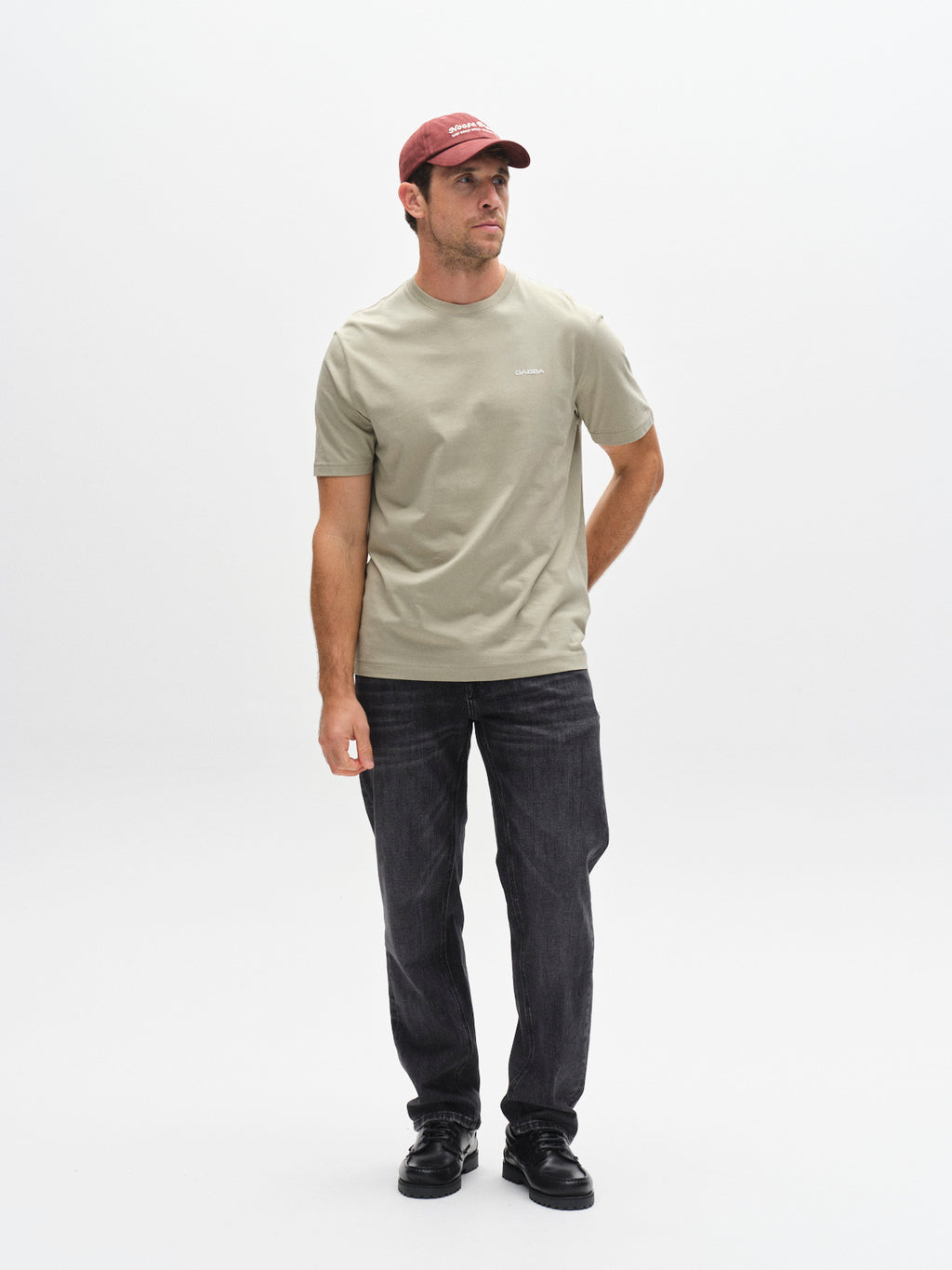 A man in a Gabba GABdune Logo GOTS SS T-Shirt in light green, black jeans, black shoes, and a red cap stands against a white background, gazing slightly to the side with one hand behind his back.