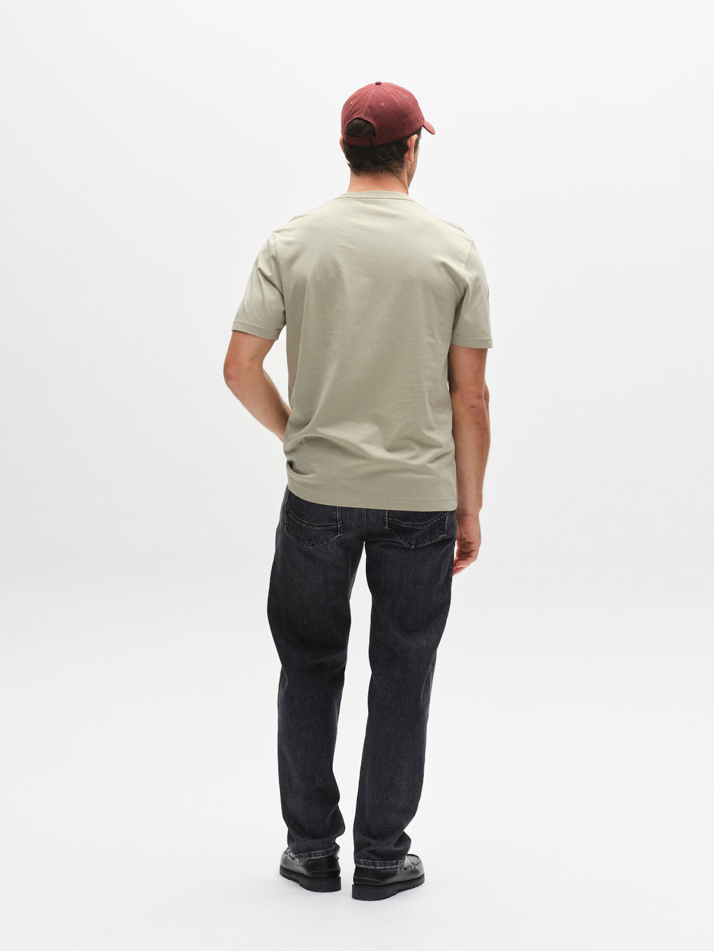 A man faces away from the camera, wearing a Gabba GABdune Logo GOTS SS T-Shirt in light green, dark jeans, black shoes, and a maroon baseball cap, standing against a plain white background.