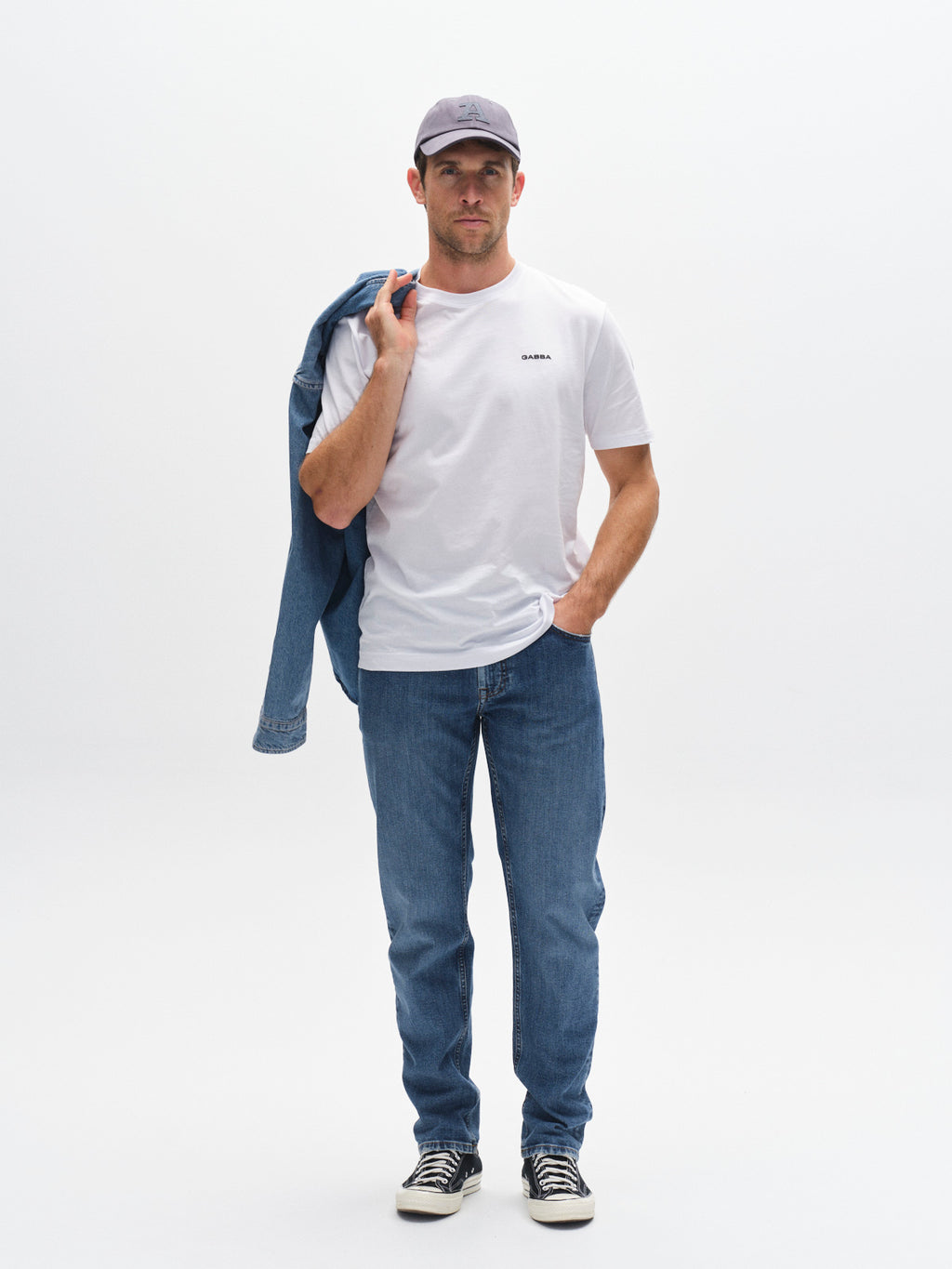 A man faces forward against a plain white background, wearing a white t-shirt, Gabba GABmarc F1010 Dark Blue Denim jeans, sneakers, and a gray cap. He has a blue jacket slung over his shoulder.
