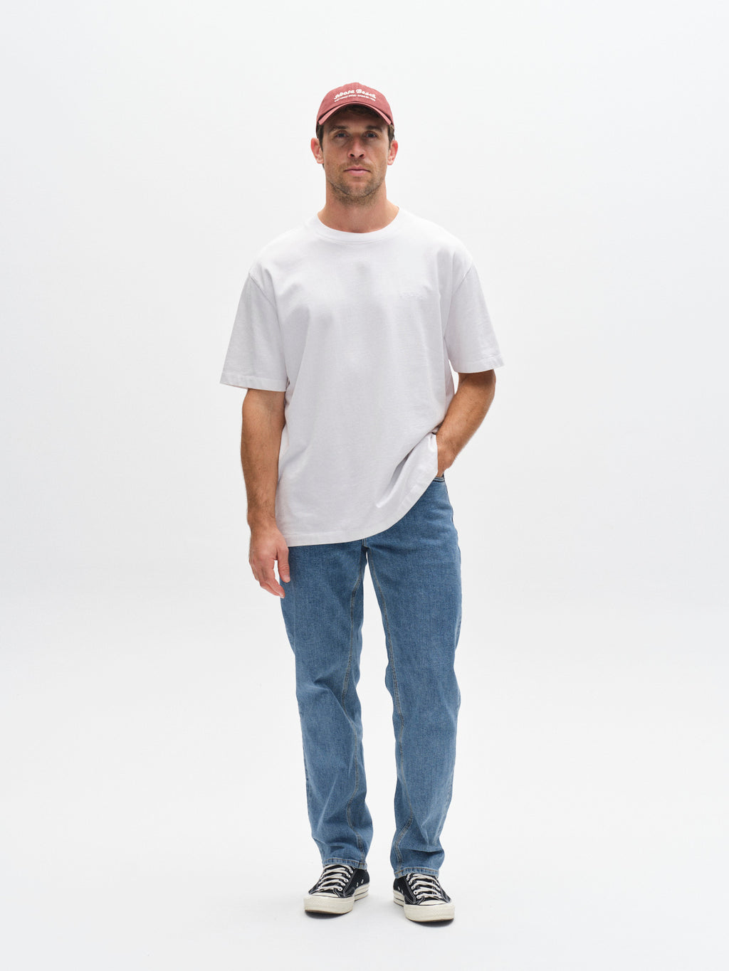 A man stands against a plain white background wearing the Gabba GABnigel Wix Boxy Hour SS T-shirt in white, vintage indigo blue jeans, a red cap, and black-and-white sneakers. One hand is in his pocket as he displays a neutral expression.