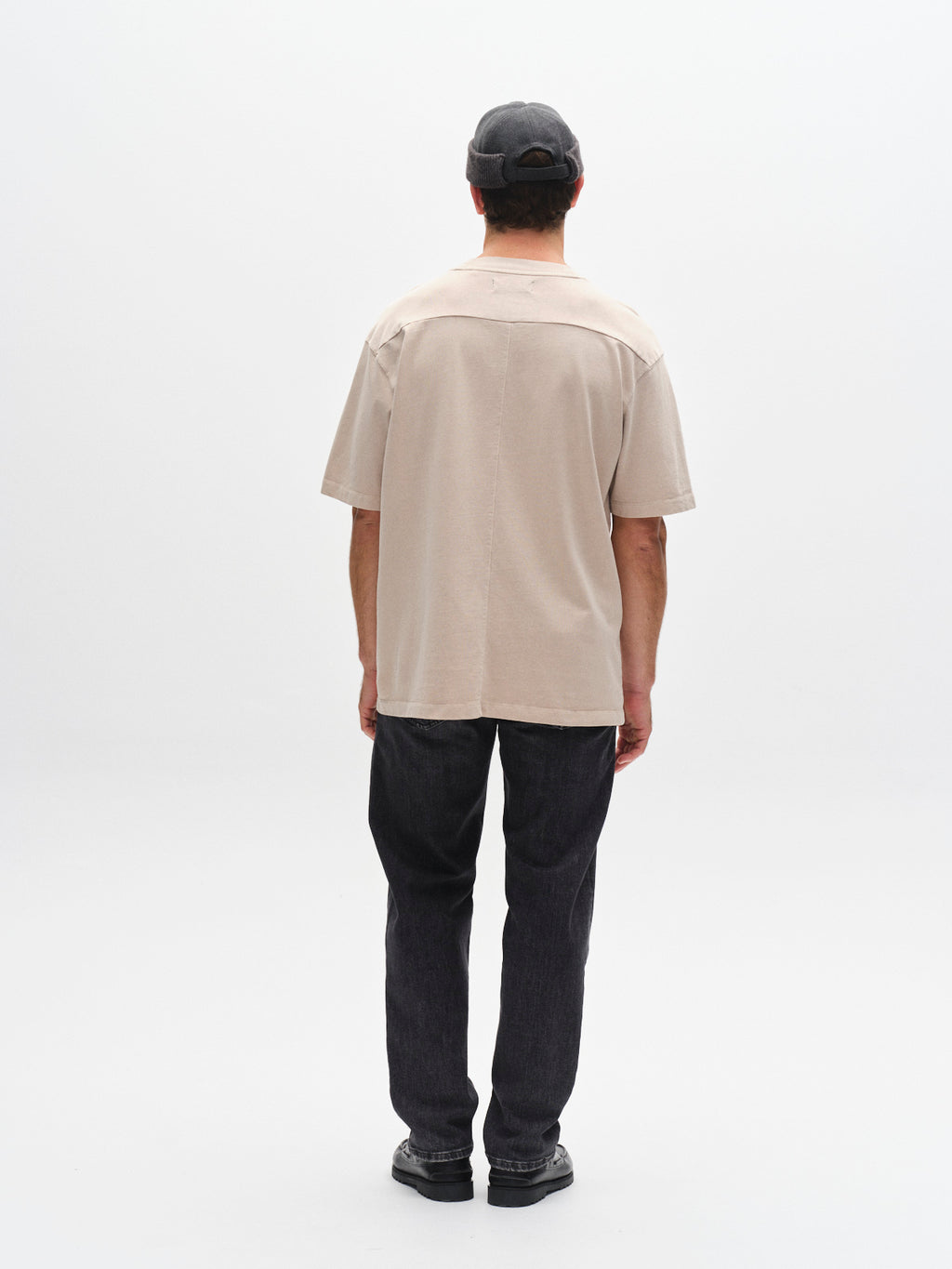 A person wearing the Gabba GABnigel Wix Boxy Hour SS t-shirt in Sand Cashmere stands with their back to the camera, paired with dark pants, black shoes, and a dark cap against a plain white background.