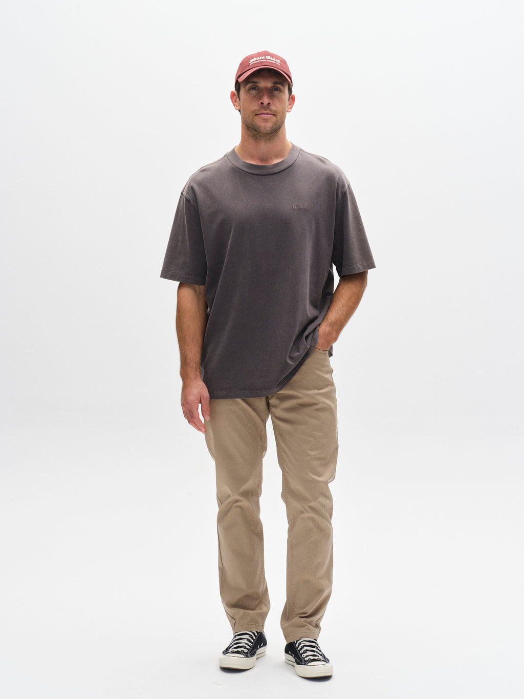 A man stands facing forward against a plain white background, wearing a red cap, beige pants, black-and-white sneakers, and the Gabba GABnigel Wix Boxy Hour SS T-shirt in dark grey. One hand is in his pocket and the other hangs by his side.