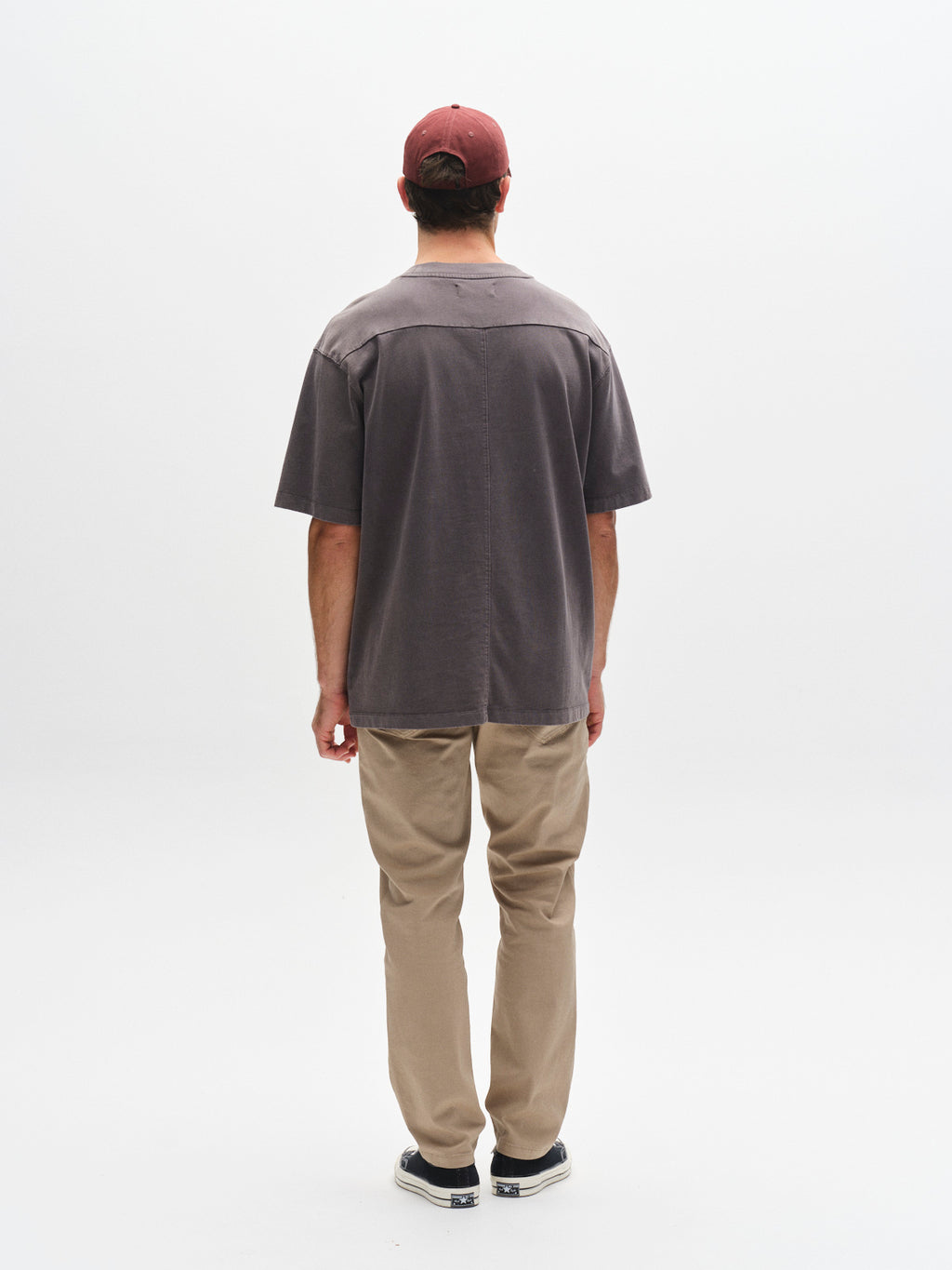 A person faces away from the camera, wearing a red cap, the Gabba GABnigel Wix Boxy Hour SS t-shirt in dark grey, beige pants, and black shoes against a plain white background.
