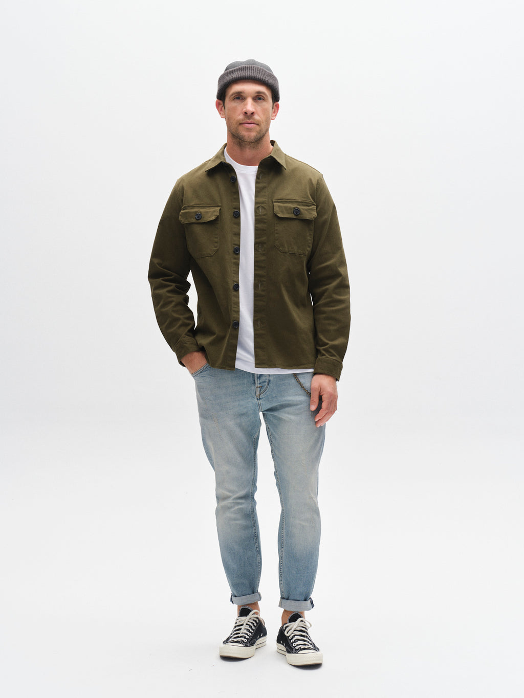 A man stands against a white background with one hand in his pocket, wearing Gabba’s GABalex Cros Cropped Lt. Blue Denim jeans, a gray beanie, green button-up over a white tee, and black sneakers.