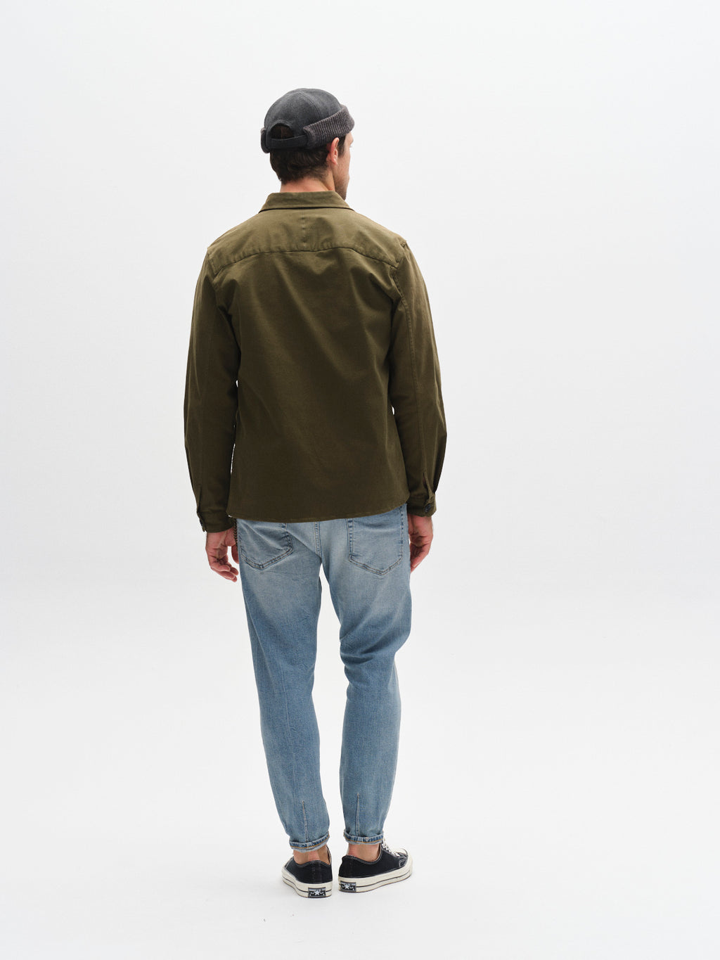 A man faces away from the camera, wearing a dark olive-green jacket, Gabba GABalex Cros Cropped - Lt. Blue Denim jeans, black slip-on shoes, and a gray flat cap against a plain light background.