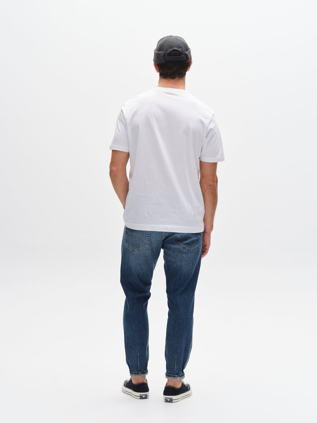 A man faces away from the camera, wearing a gray cap, plain white tee, black slip-on shoes, and Gabba GABalex Cros Cropped jeans in mid blue denim against a white background.