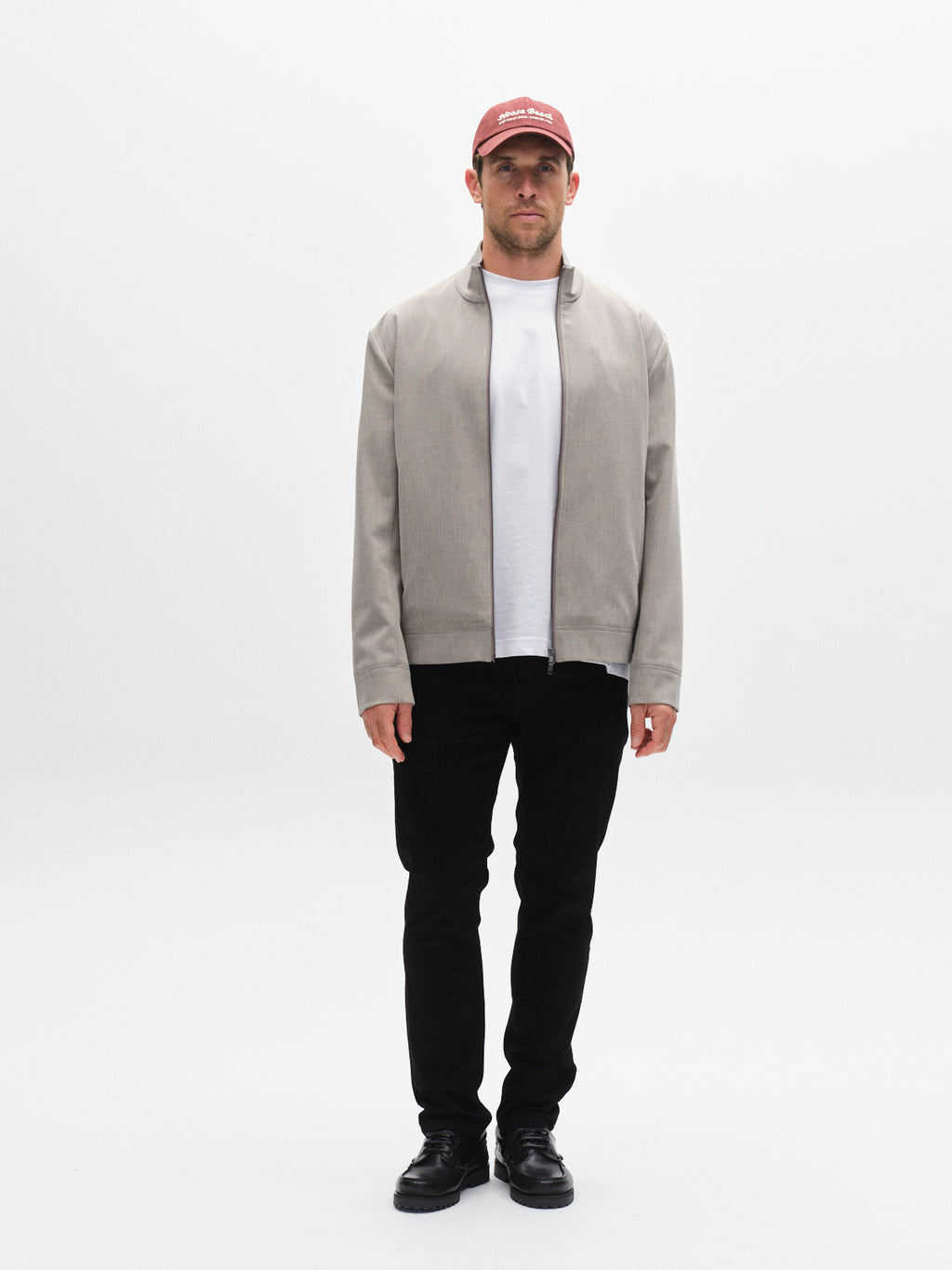 A man faces forward against a plain white background, wearing a red baseball cap, light gray jacket over a white t-shirt, Gabba GABmarc Sword - Black Denim slim fit pants, and black shoes for a sharp, clean look.