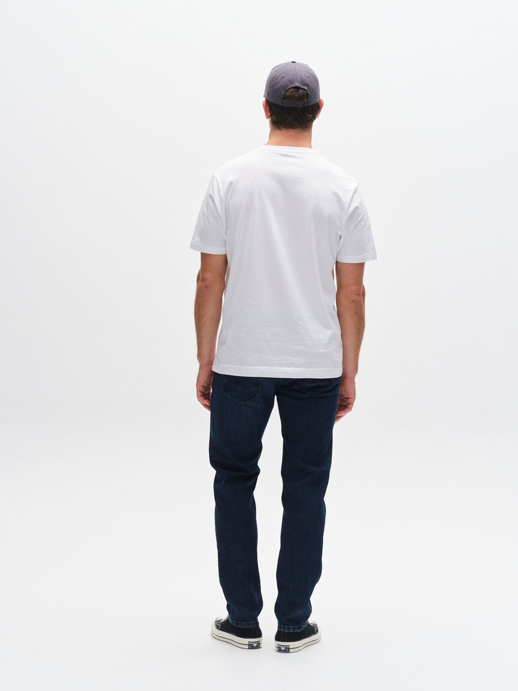 A man faces away from the camera, wearing a gray cap, white tee, black sneakers, and Gabba GABmarc Mego - Dark Blue Denim jeans crafted with Wiser Wash technology, all set against a plain white background.