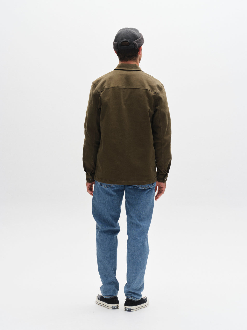 A person stands facing away from the camera, wearing a dark cap, olive green jacket, black-and-white sneakers, and Gabba GABmax Cros - Lt. Blue Denim jeans against a plain white background.