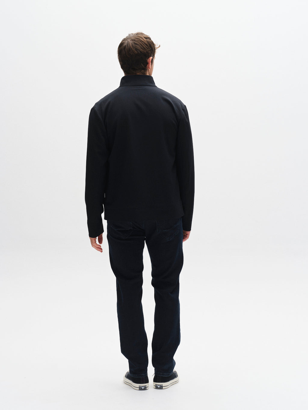 A man stands facing away from the camera against a plain white background, wearing a black jacket, Gabba GABmarc Panter - Dark Blue Denim jeans, and sneakers.