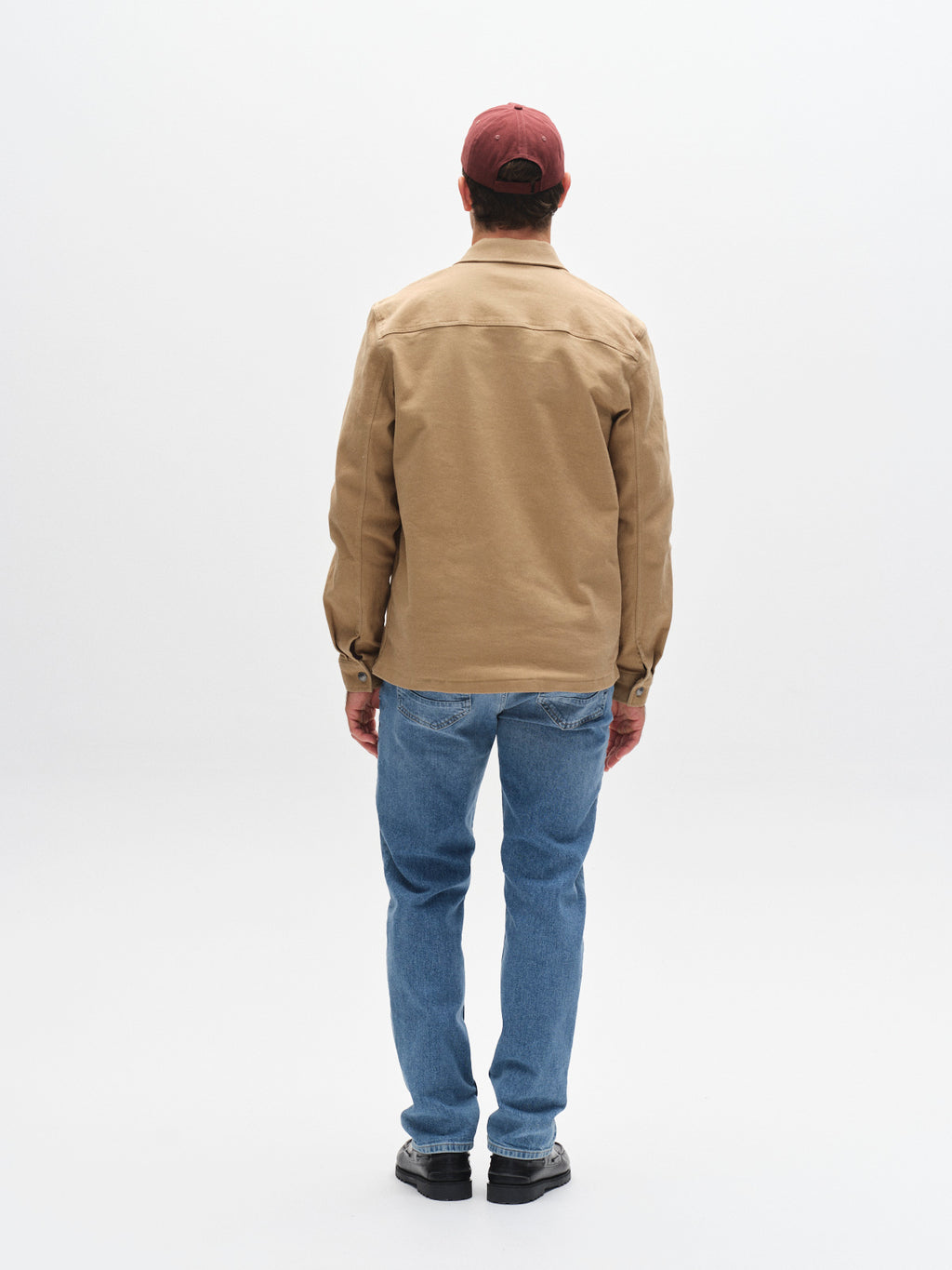A man stands facing away from the camera in a tan jacket, black shoes, a red baseball cap, and Gabba GABMarc 5poc - Mid Blue Denim jeans against a plain white background.