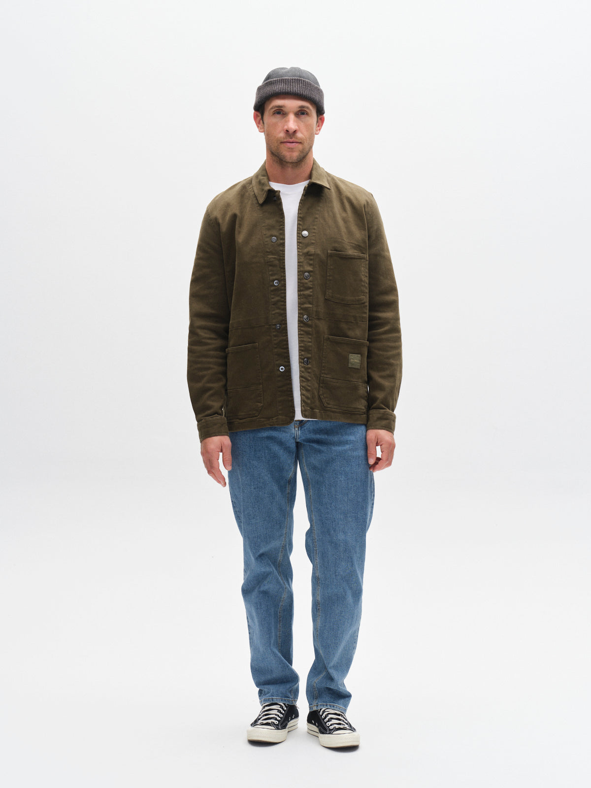 A man stands facing forward against a plain white background, wearing a gray beanie, Gabba GABbeta Twill - Army overshirt, light blue jeans, and black sneakers.