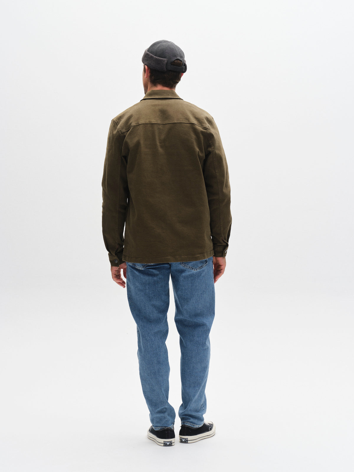 A person stands facing away, wearing a Gabba GABbeta Twill - Army overshirt, a dark cap, blue jeans, and black-and-white shoes against a plain white background.