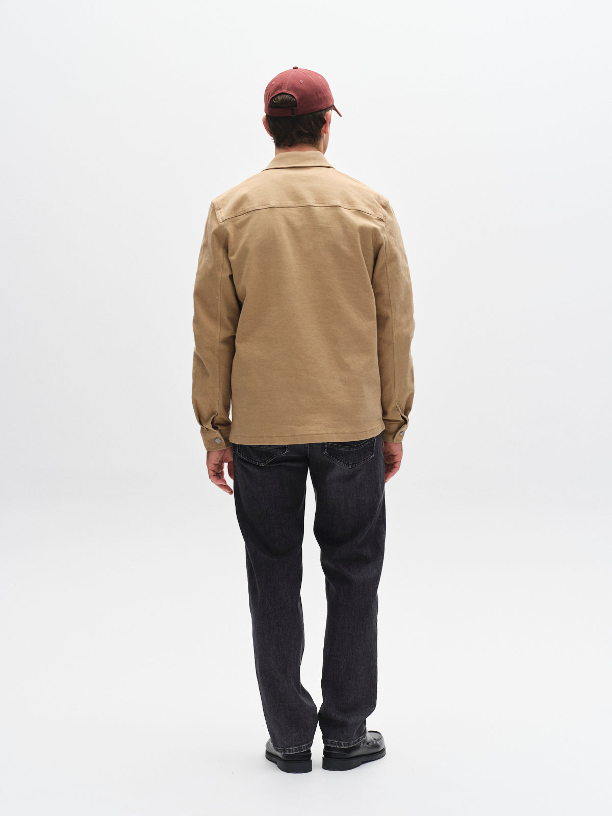 A person stands facing away from the camera, wearing a red cap, Gabba GABbeta Twill - Sand jacket, dark jeans, and black shoes against a plain white background.