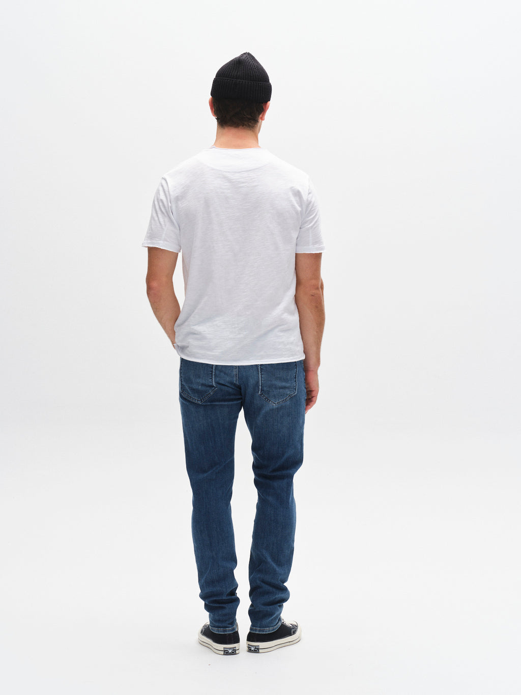 A man faces away from the camera, wearing a black beanie, plain white t-shirt, Gabba GABjones Vanew - Mid Blue Denim jeans with a slim fit and high flexibility, and sneakers, set against a plain white background.