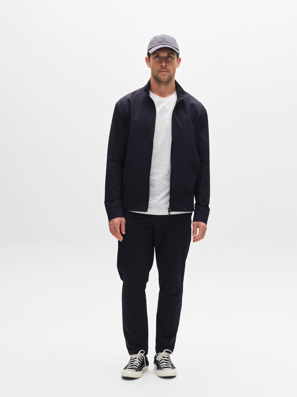 A man faces forward against a plain white background, wearing the Gabba GABnik Tech - Navy zip-front jacket, paired with a gray cap, white T-shirt, black pants, and black-and-white sneakers.