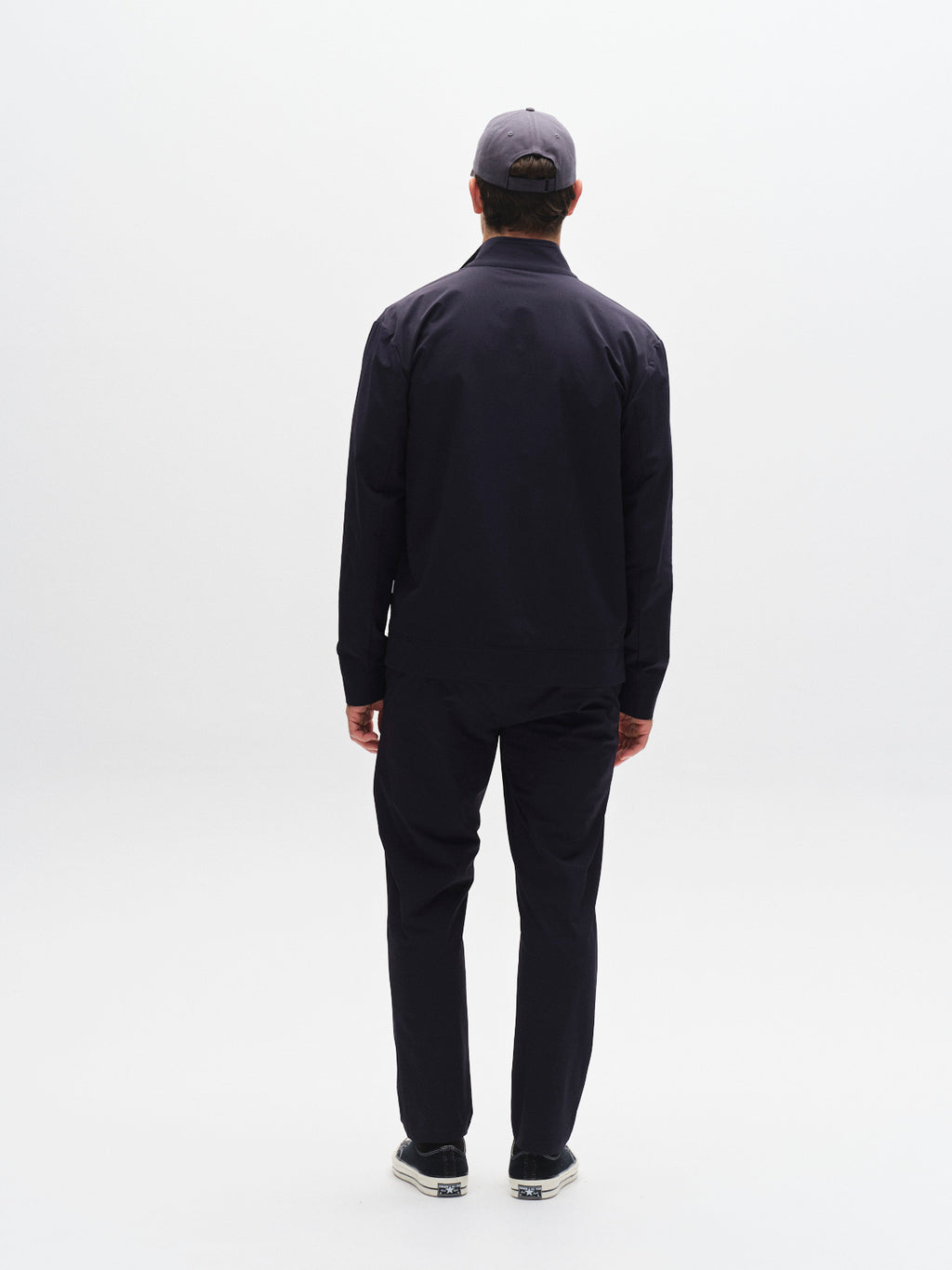 A man faces away from the camera against a plain white background, wearing the Gabba GABnik Tech - Navy jacket, dark pants, dark shoes, and a gray cap.
