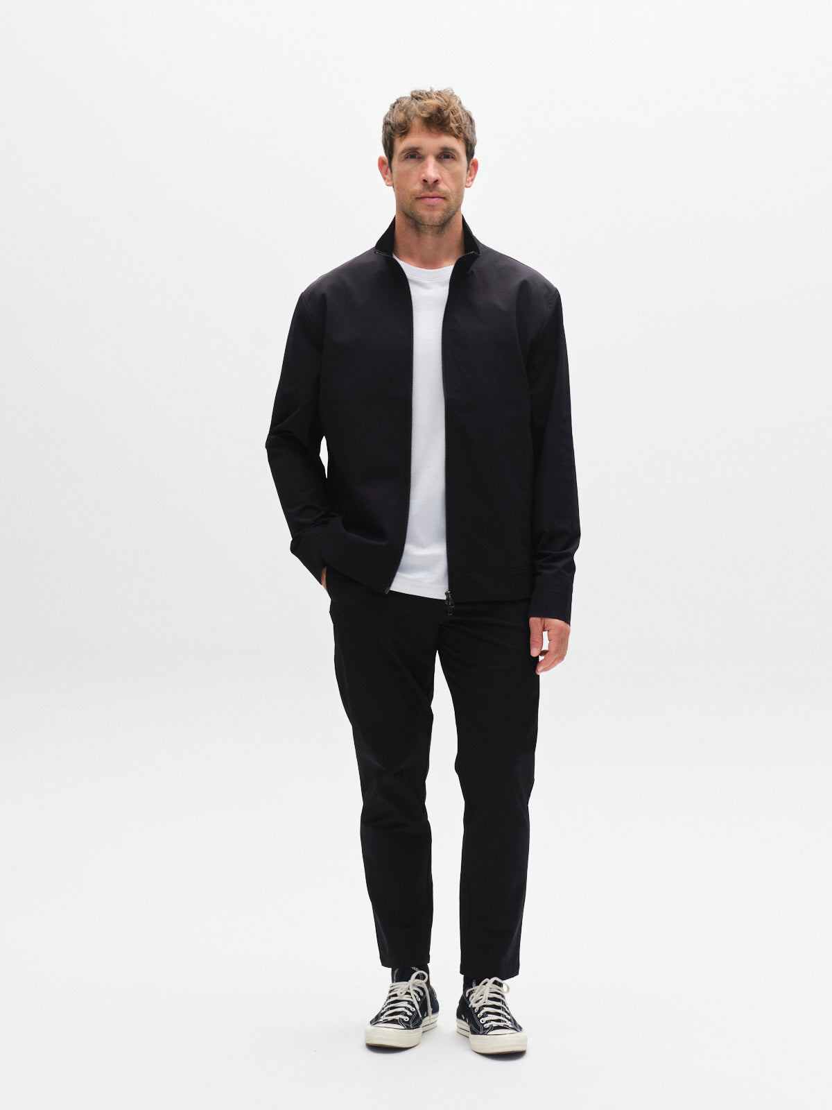 A man stands against a white background wearing the Gabba GABnik Tech - Black jacket, paired with a white t-shirt, black pants, and black sneakers with white laces. His left hand is in his pocket as he looks straight ahead.