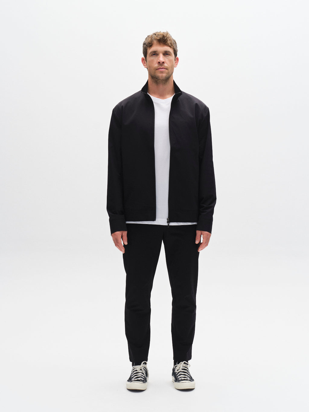 A man faces forward against a plain white background, wearing a black jacket, white t-shirt, Gabba GABmonza Tech - Black relaxed straight fit pants, and black-and-white sneakers.