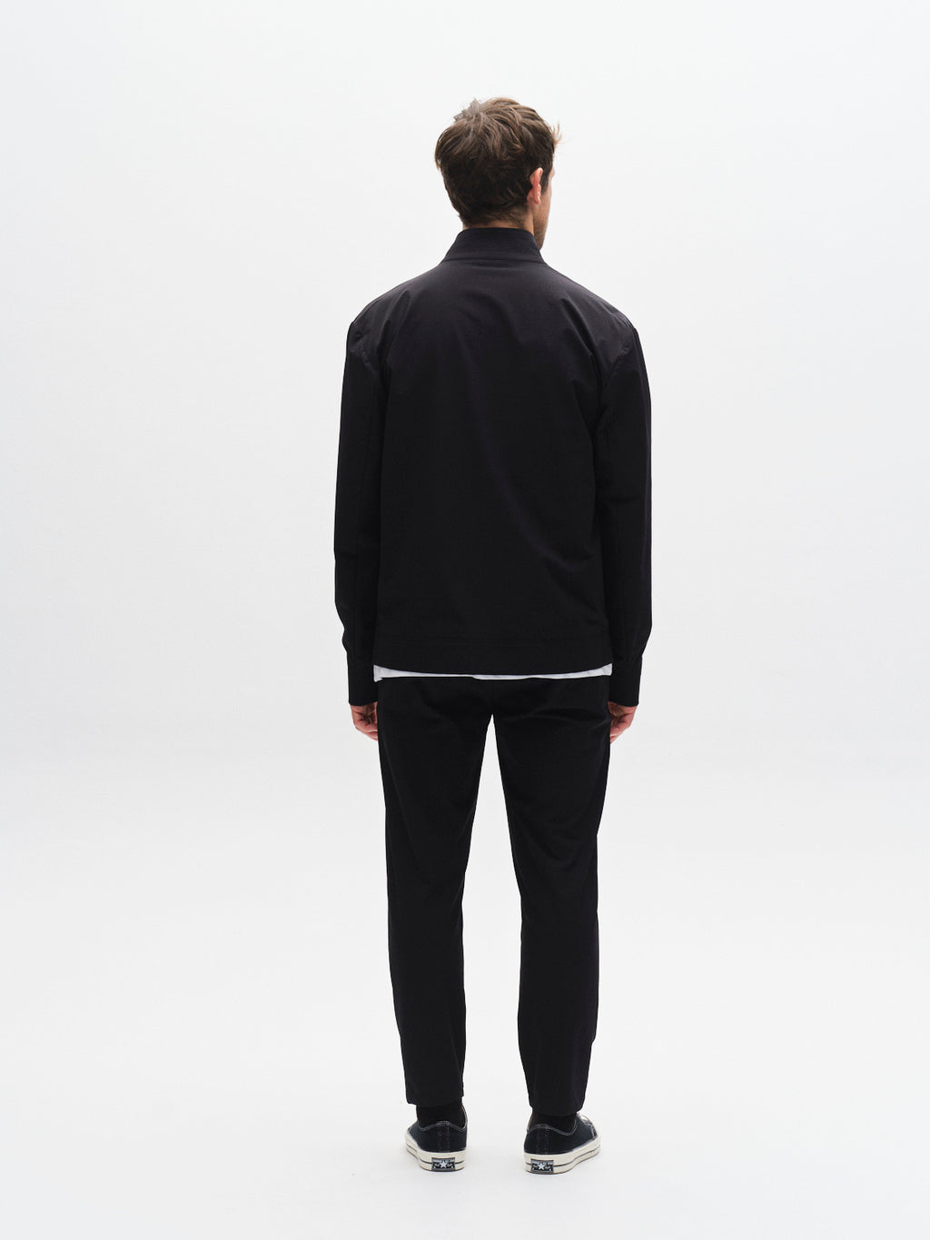 A man stands facing away in a minimal white studio wearing the Gabba GABmonza Tech - Black jacket, relaxed straight fit elastic waist pants, and black slip-on shoes with white soles.