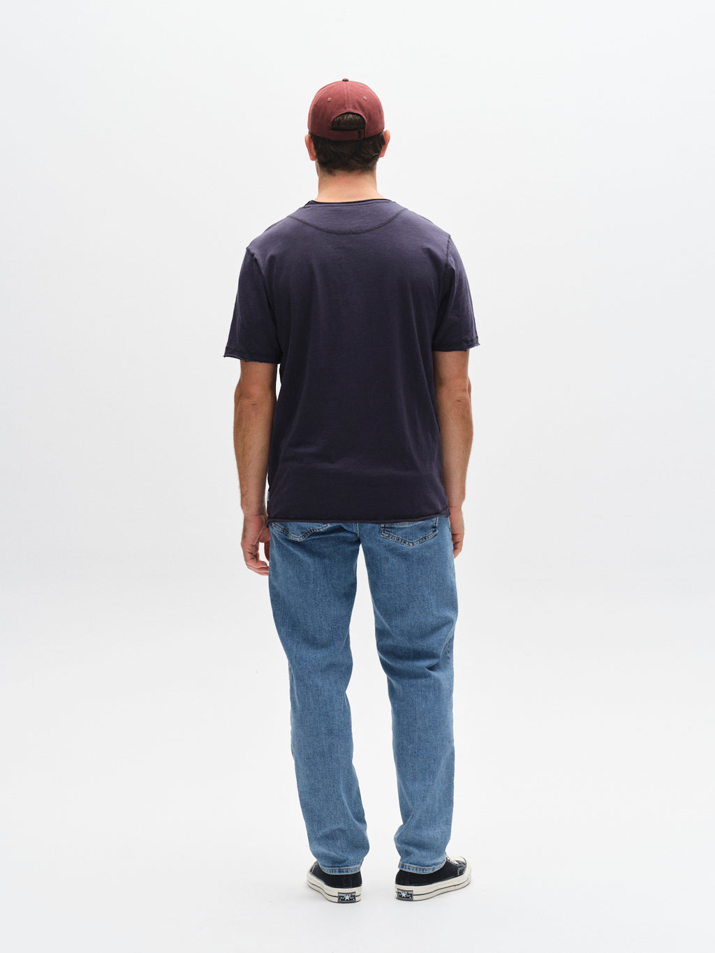 A man faces away from the camera, wearing the Gabba GABkostant New SS t-shirt in navy, paired with a maroon cap, light blue jeans, and black sneakers against a plain white background.