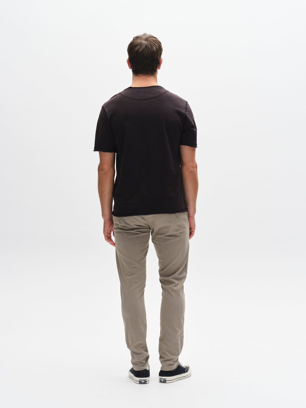 A man faces away from the camera in a white studio, wearing the Gabba GABkostant New SS - Meteorite dark slub t-shirt, light khaki pants, and black sneakers. The background is clean and uncluttered.