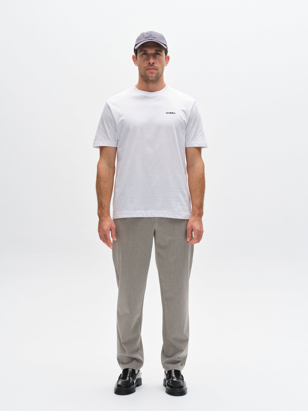 A man stands facing forward, wearing a light gray cap, a plain white t-shirt, Gabba GABnapoli 19 Phin GRS trousers in sand, and black shoes, against a plain white background.