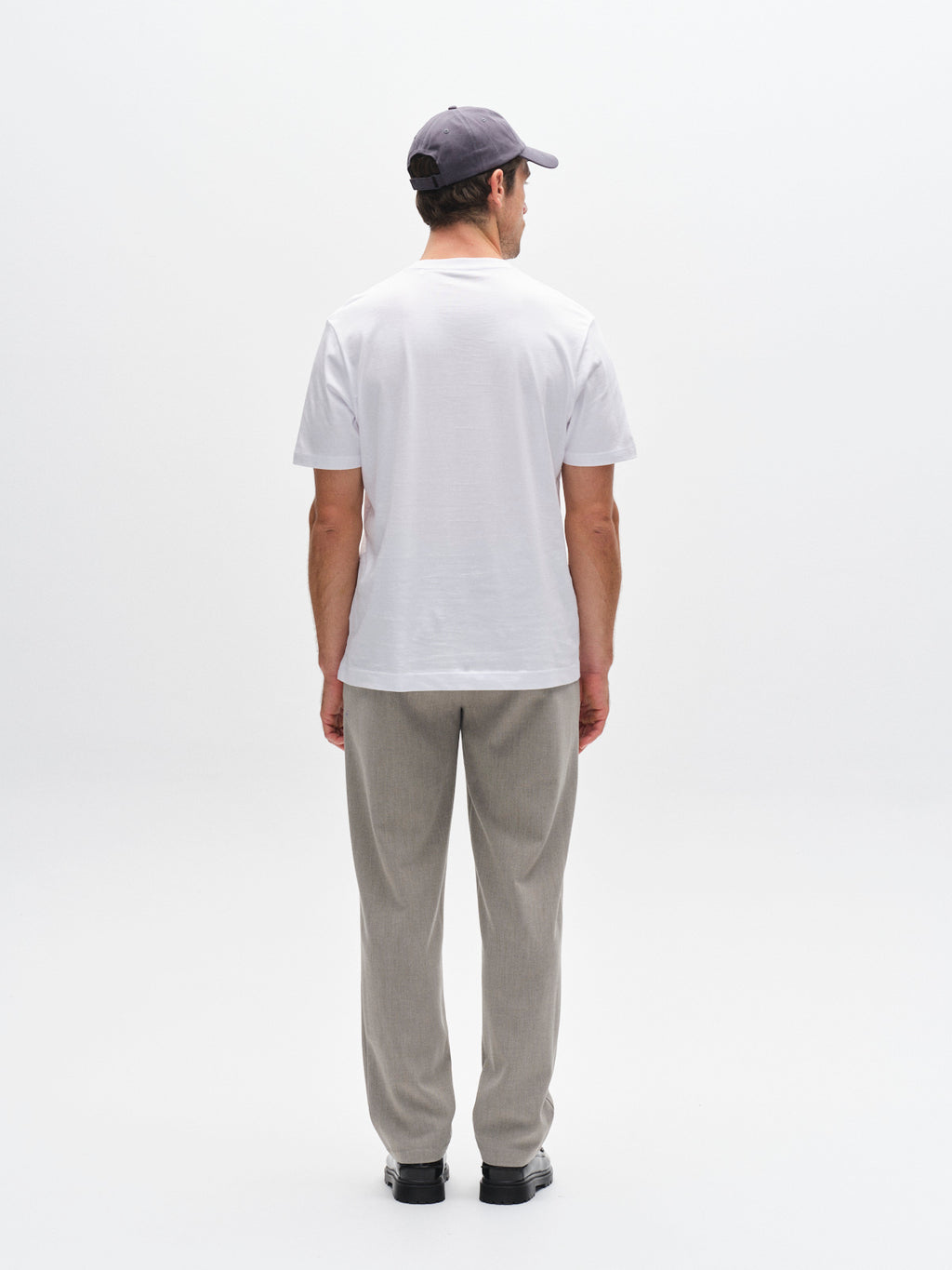 A man stands facing away, wearing a white t-shirt, GABnapoli 19 Phin GRS - Sand wool blend chino pants by Gabba in a regular straight fit, black shoes, and a dark baseball cap, against a plain white background.