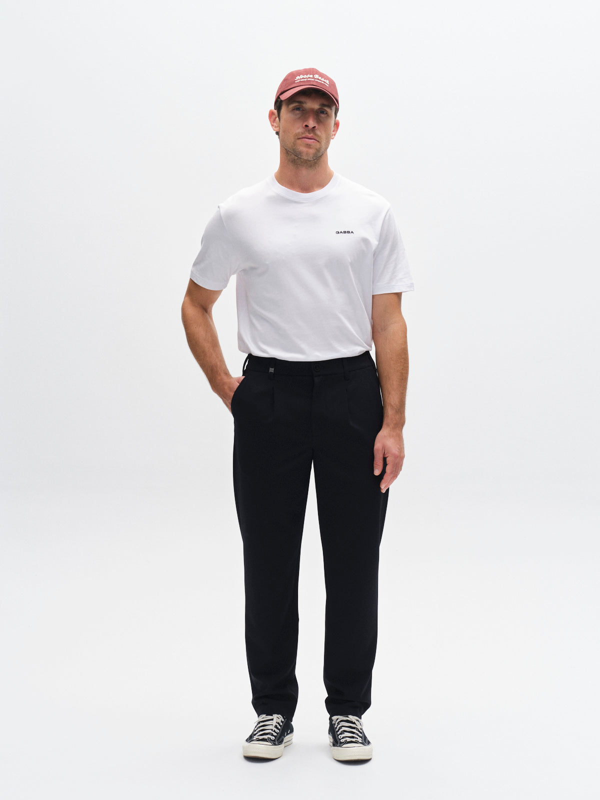 A man faces forward in a studio, wearing a red cap, white T-shirt, Gabba GABnapoli 19 Phin GRS - Blue Black straight fit chinos, and black sneakers. One hand is in his pocket against a plain white background.