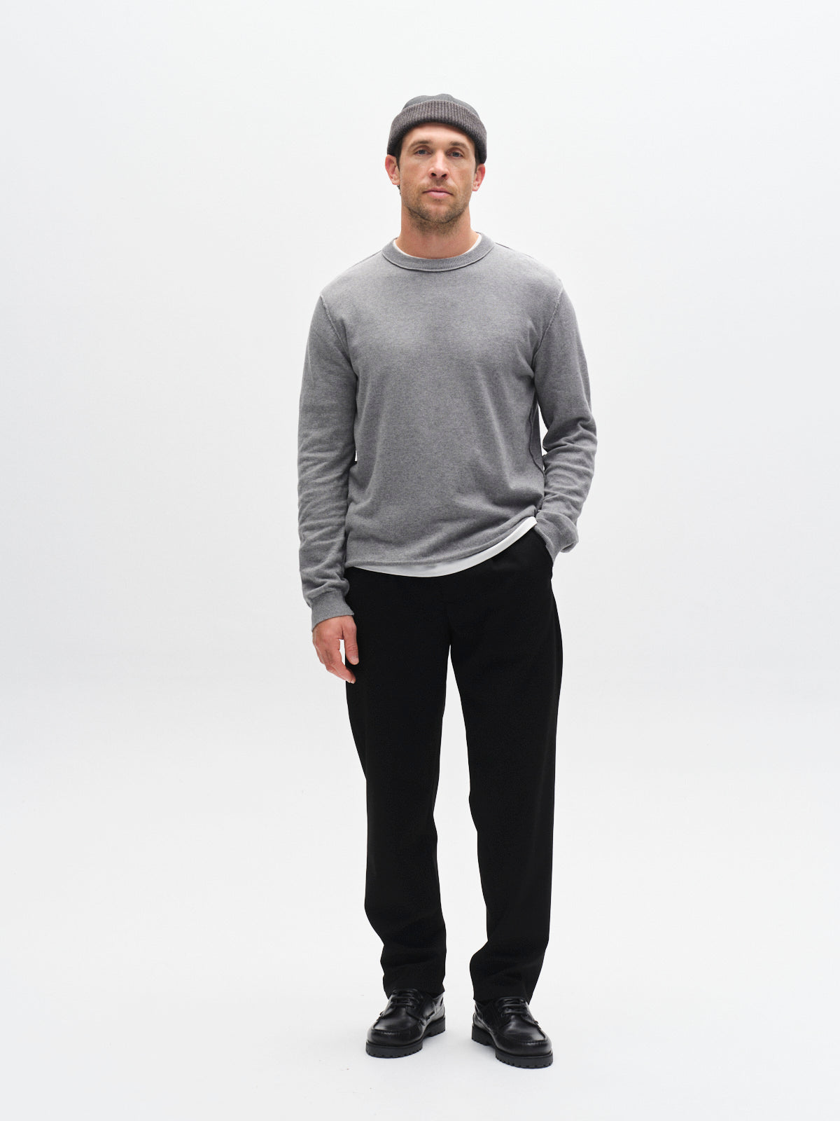 A man wearing a gray beanie, gray long-sleeve shirt, black Gabba GABnapoli 19 Phin GRS tailored chinos in a regular straight fit, and black shoes stands against a plain white background with one hand in his pocket.