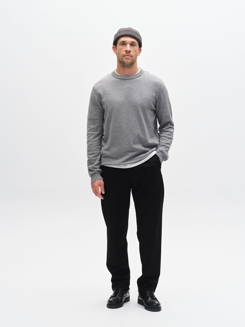 A man stands against a plain white background, wearing Gabba's GABnapoli 19 Phin GRS - Black pants with a gray sweater, black shoes, and a gray knit beanie. One hand is in his pocket as he looks forward with a neutral expression.