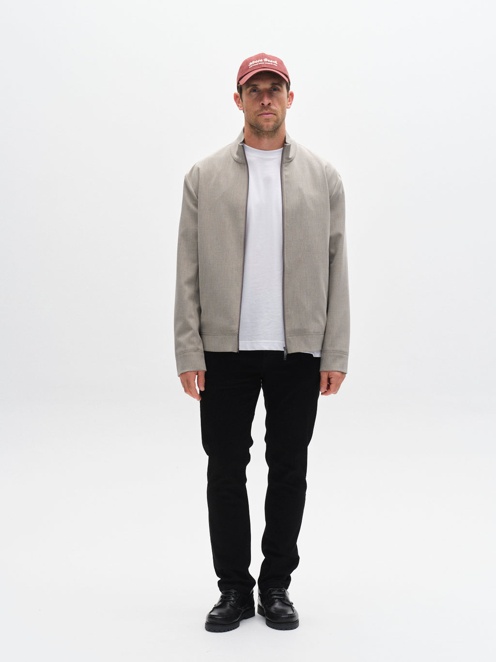 A man faces forward against a plain white background, wearing the Gabba GABnik Phin GRS - Sand jacket made from recycled polyester, paired with a white t-shirt, black pants, black shoes, and a red baseball cap.
