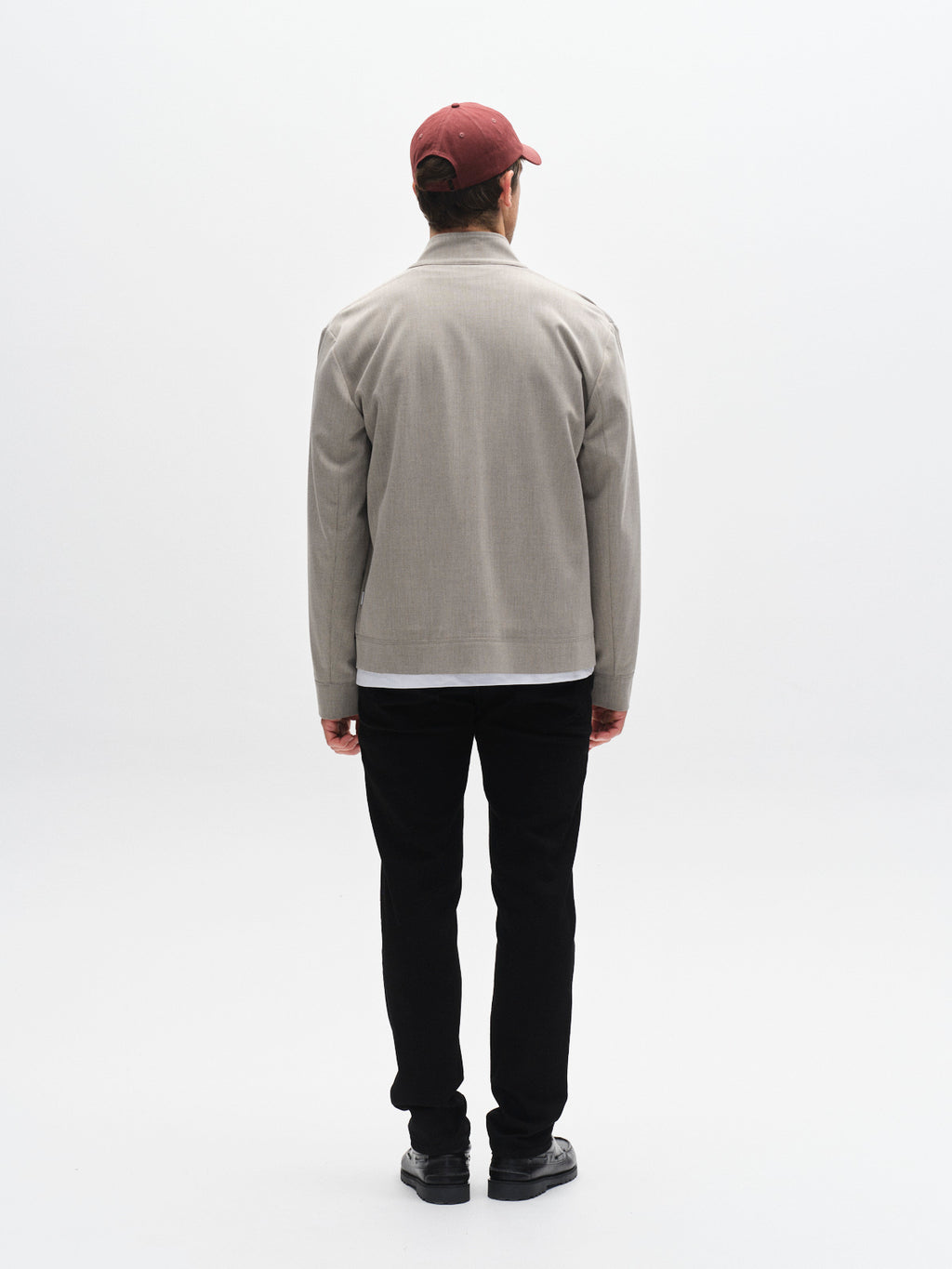 A man stands in a plain, light studio wearing the Gabba GABnik Phin GRS - Sand beige tailored jacket (made from GRS-recycled polyester), black pants, black shoes, and a red baseball cap. His arms hang relaxed at his sides.