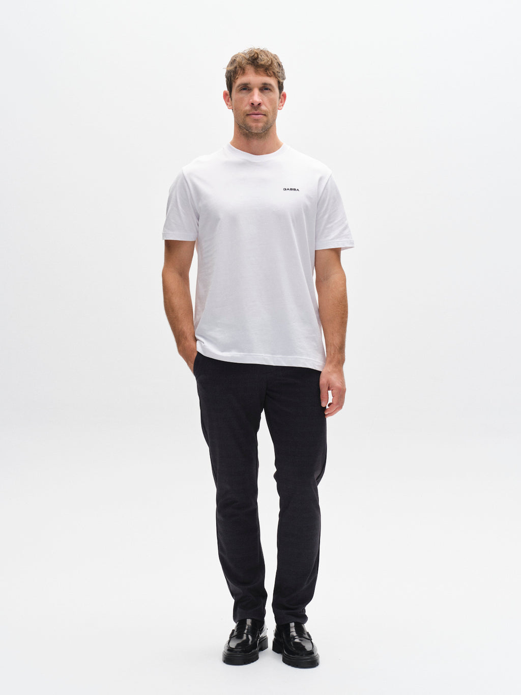 A man stands facing forward against a plain white background, wearing the Gabba GABpaul Nygo - Navy slim straight fit trousers made from comfort stretch fabric, paired with a white short-sleeve t-shirt and black shoes.