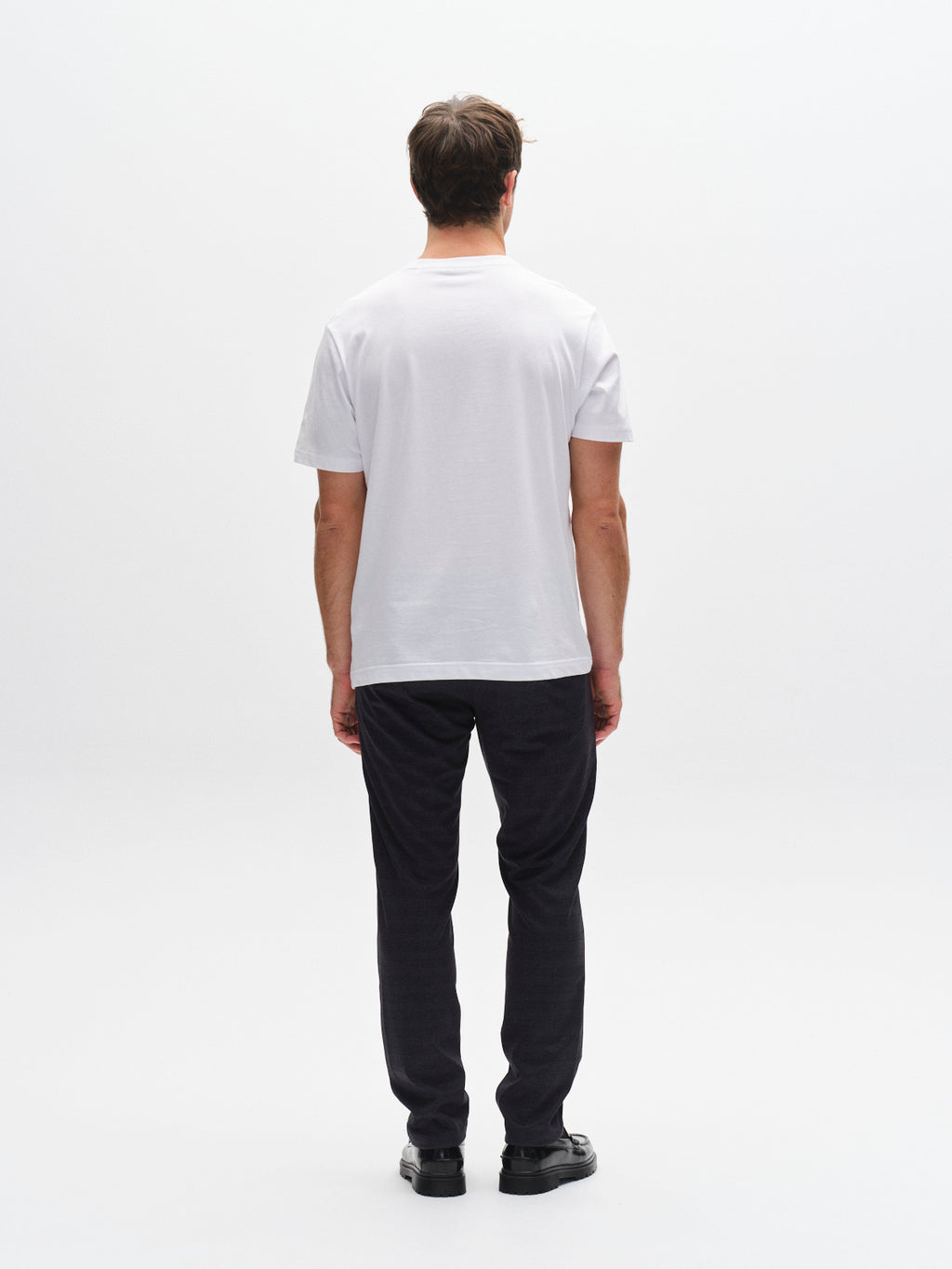 A man, seen from behind against a white background, wears Gabba’s GABpaul Nygo - Navy slim straight fit trousers in dark stretch fabric with a plain white t-shirt and black shoes.