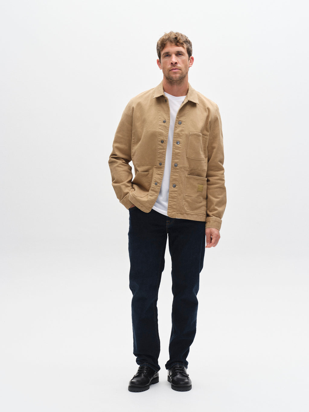 A man stands against a plain white background, wearing a tan button-up jacket over a white t-shirt, GABmarc Jingo - Dark Blue Denim jeans by Gabba crafted from comfort stretch denim in a straight fit, and black shoes.