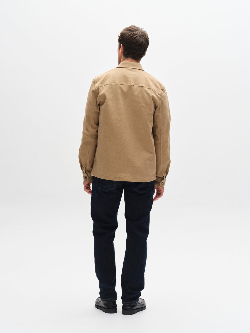 A man stands facing away from the camera against a plain white background, wearing a tan jacket, black shoes, and straight fit Gabba GABmarc Jingo dark blue denim jeans crafted from comfort stretch fabric.