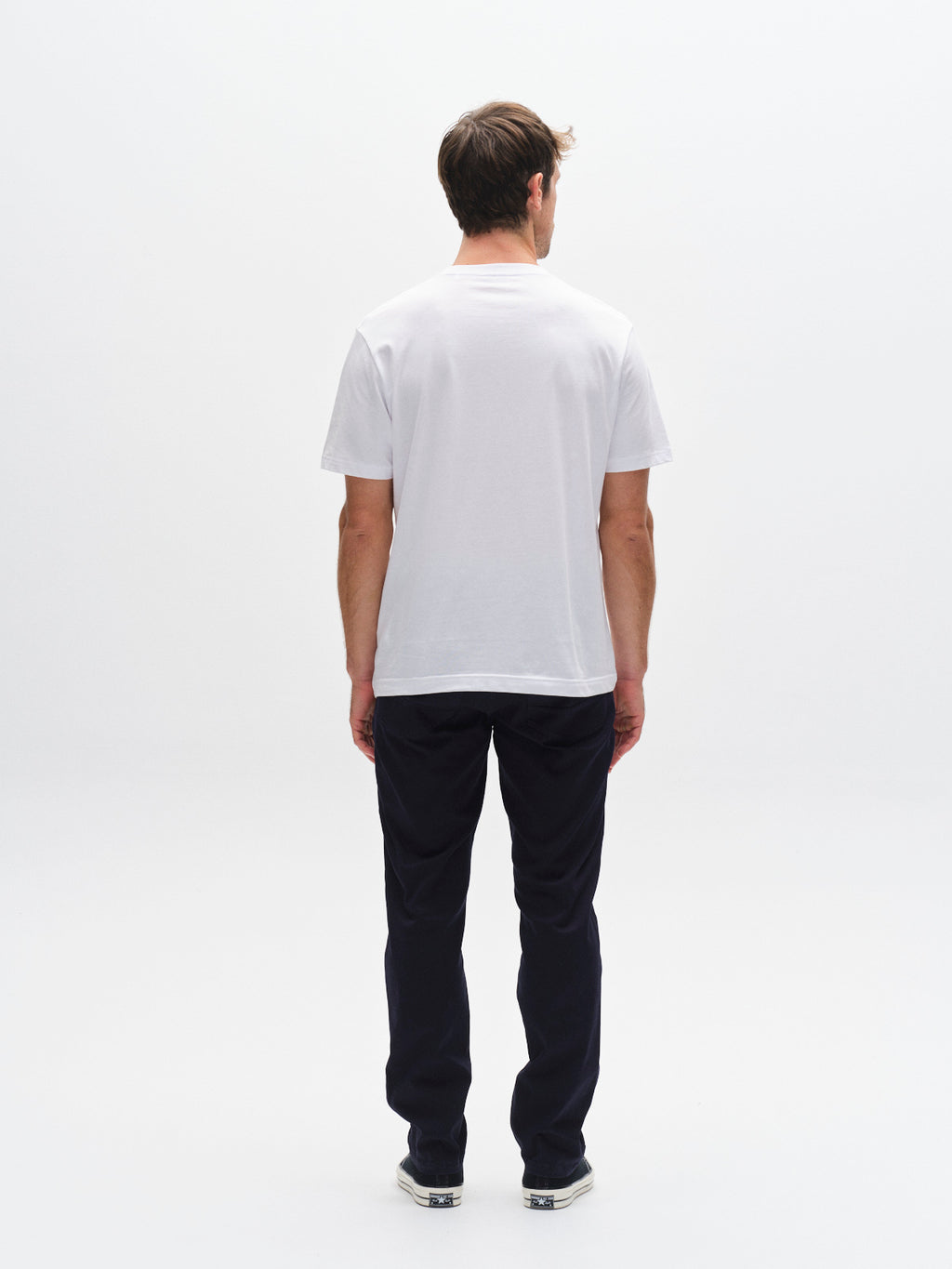 A man faces away from the camera against a white background, wearing Gabba’s GABmarc 5poc Marlo jeans in Dark Navy, paired with a plain white t-shirt and sneakers.