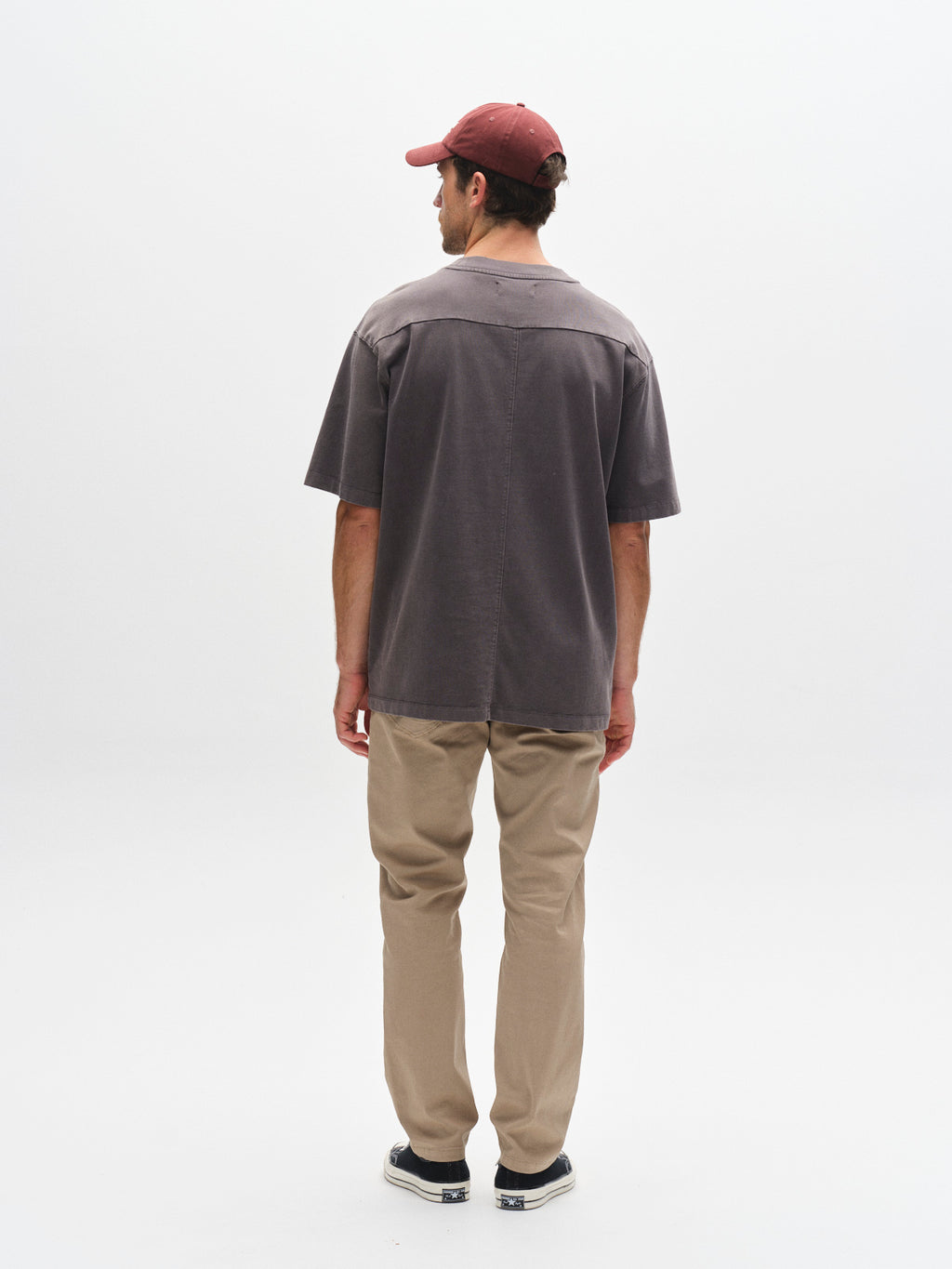 A man faces away from the camera wearing Gabba GABmarc 5poc Marlo - Coriander jeans, a maroon cap, loose gray T-shirt, and black slip-on shoes against a white background—ideal for everyday style.