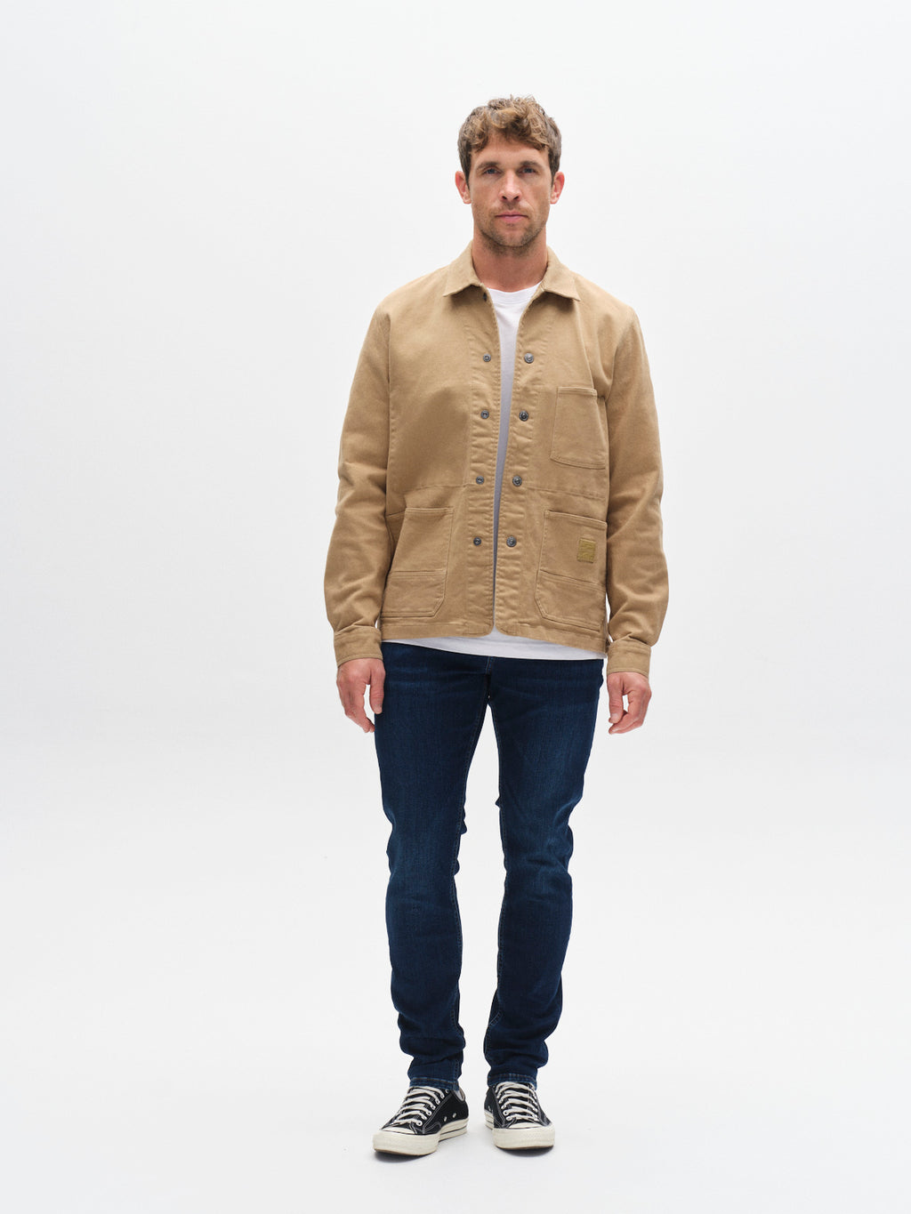 A man stands facing forward wearing a tan jacket, white shirt, and Gabba GABjones Dhera Jeans in mid blue denim—a slim straight fit style crafted from superstretch fabric—paired with black sneakers with white soles, against a plain white background.