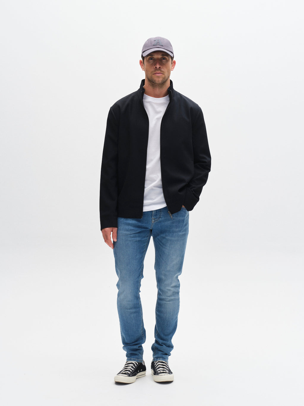A man wearing a gray cap, black jacket, white t-shirt, and Gabba GABjones Dhera Jeans in light blue denim stands facing forward against a plain white background, paired with black sneakers.