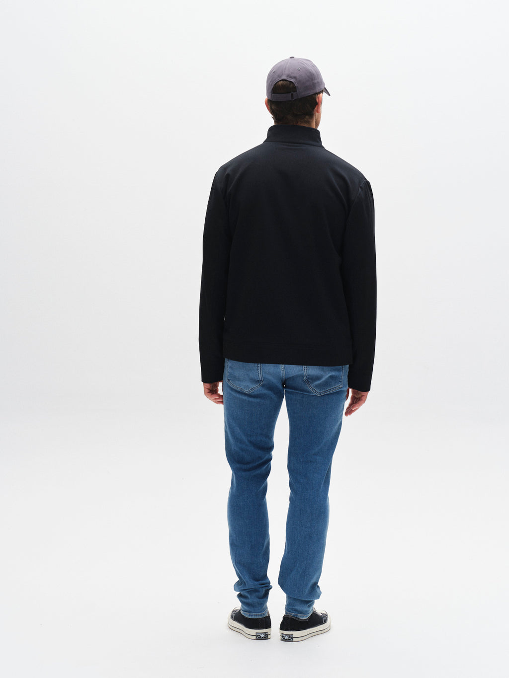 A man wearing a black jacket, Gabba GABjones Dhera Jeans in light blue denim, black sneakers, and a gray cap stands facing away from the camera against a plain white background.