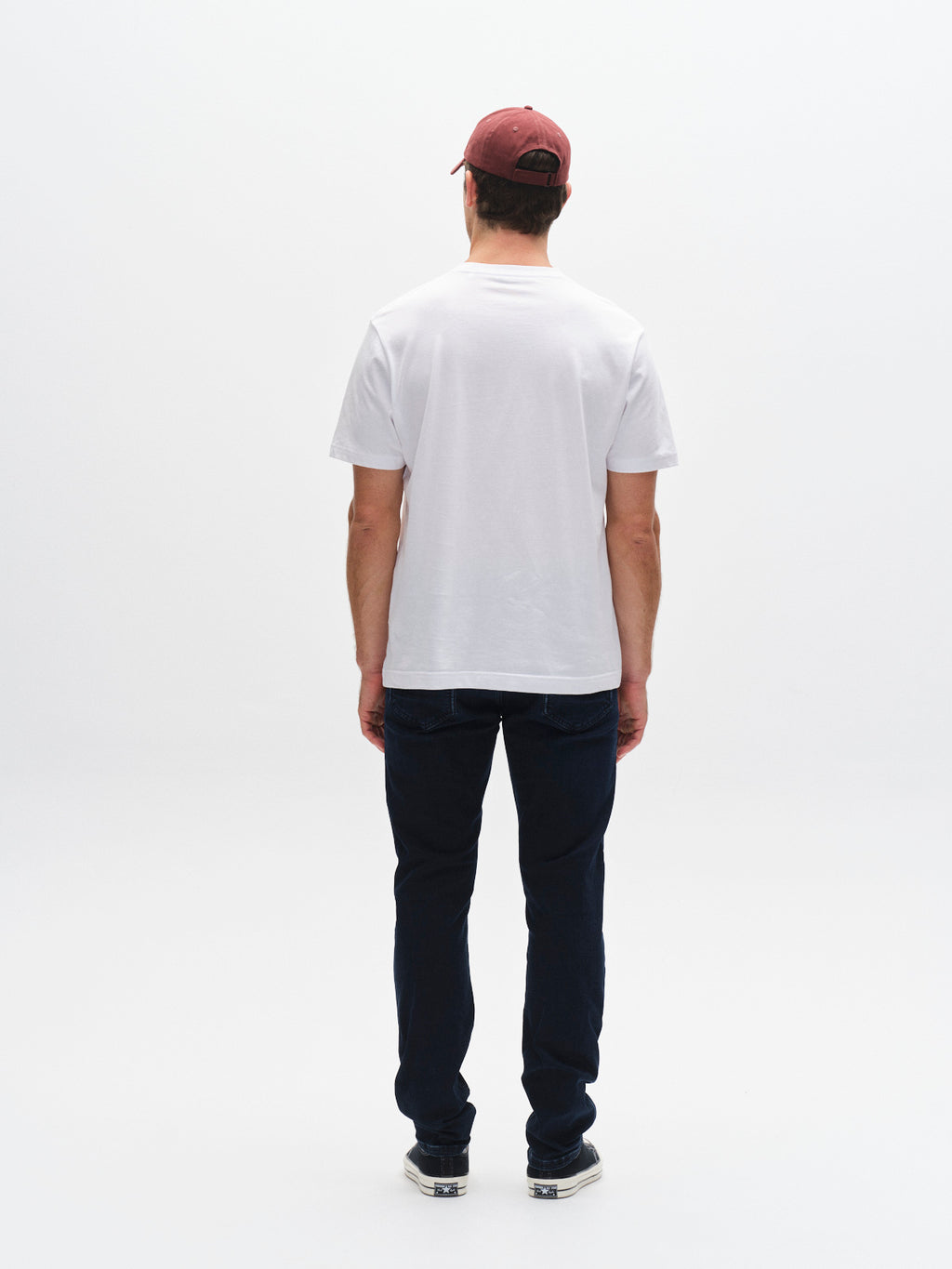 A man stands with his back to the camera, wearing a red cap, plain white t-shirt, Gabba GABjones Blueblack Jeans in blue black denim for flexibility and comfort, and sneakers, against a plain white background.