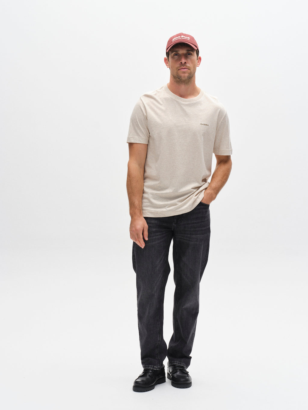 A man wearing a Gabba GABdune Melange New SS T-Shirt in Sand Mel., dark jeans, black shoes, and a red cap stands against a plain white background with one hand in his pocket.