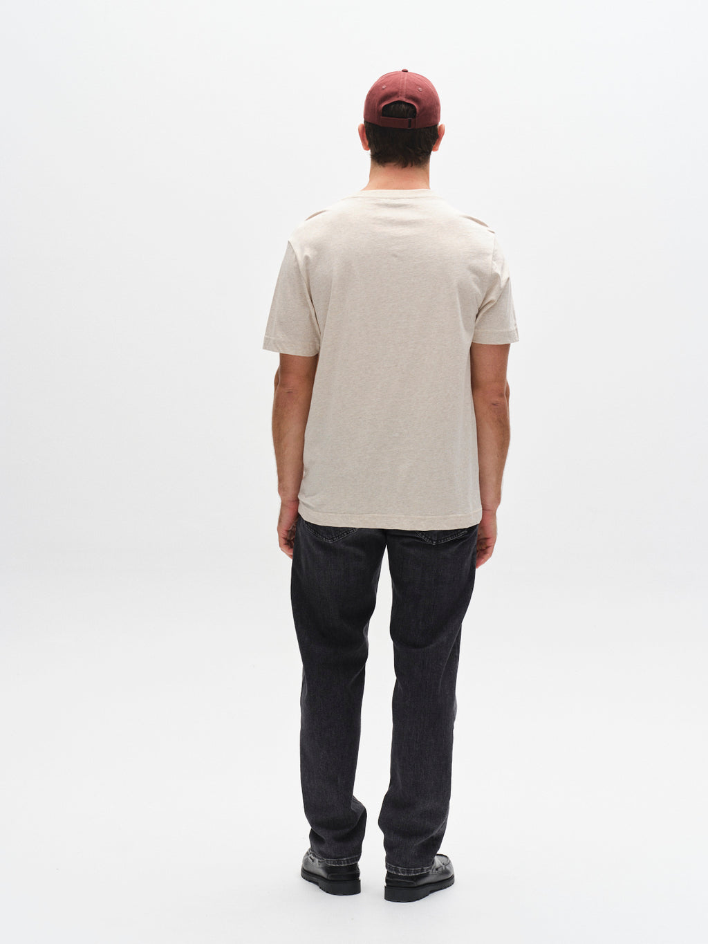 A person wearing a Gabba GABdune Melange New SS T-Shirt in Sand Mel., dark jeans, black shoes, and a red baseball cap stands facing away against a plain white background.