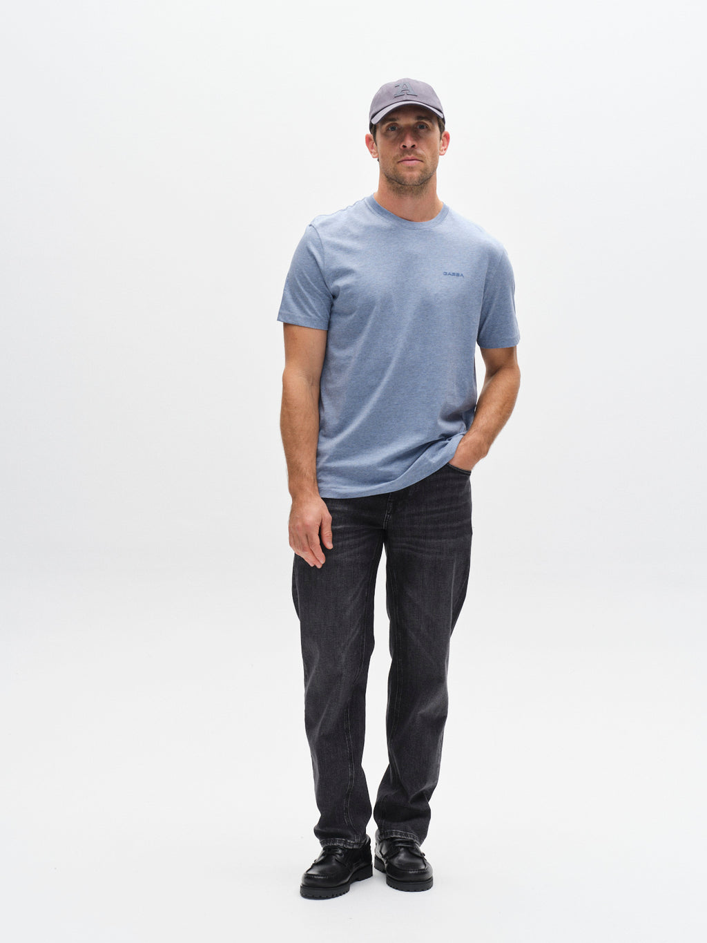 A man stands against a white background wearing the Gabba GABdune Melange New SS T-Shirt in Blue Melange, dark jeans, black shoes, and a gray baseball cap. He has one hand in his pocket and a neutral expression.