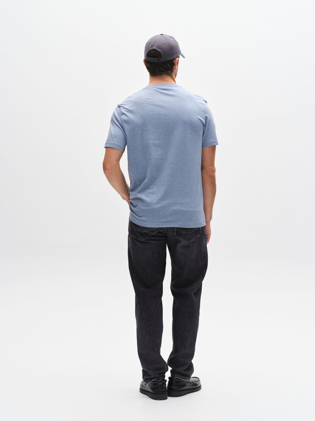 A person wearing the Gabba GABdune Melange New SS T-Shirt in Blue Melange, paired with dark jeans, black shoes, and a gray cap stands facing away from the camera against a plain white background.