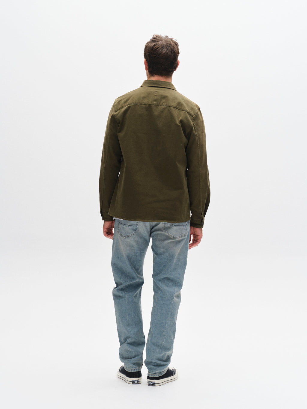 A man faces away from the camera, wearing a green long-sleeve shirt, Gabba GABcarl CrosV2 Jeans in Lt. Blue Denim, and black-and-white sneakers against a plain white background.