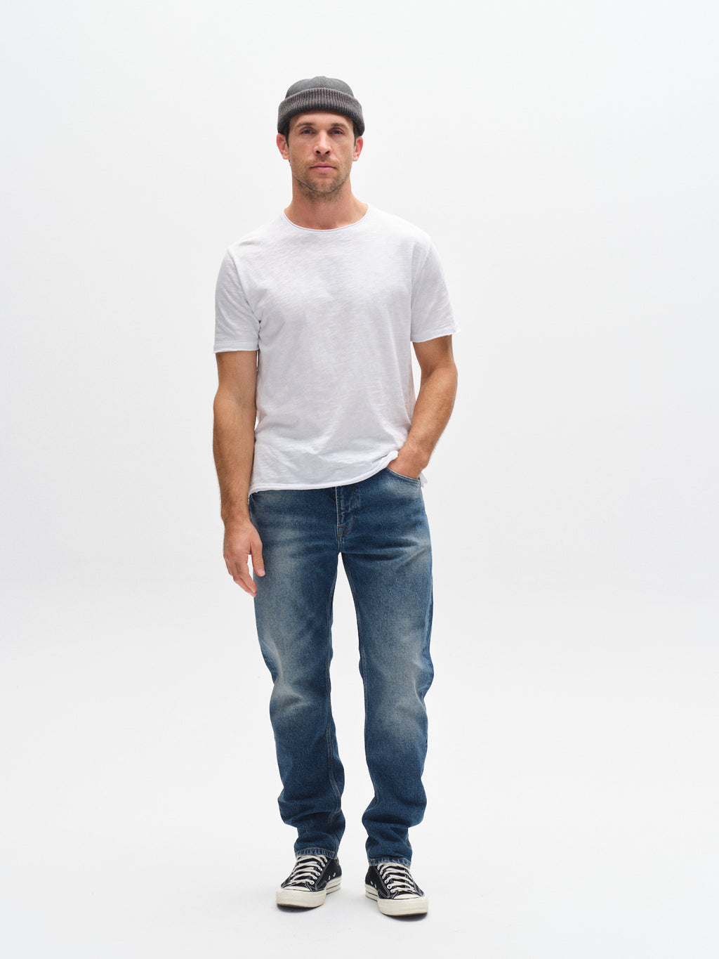A man stands against a plain white background wearing a gray beanie, white t-shirt, Gabba GABcarl OskinV2 Jeans in dark blue denim, black-and-white sneakers, and has one hand in his pocket.