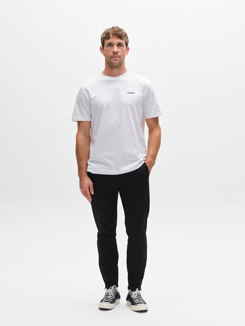 A man with short brown hair and a metro style stands against a white background wearing a white t-shirt, black GABBA Pisa Small Dot pants in high-stretch cotton-blend, and black sneakers with white laces, his left hand in his pocket.